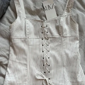 Vit randig korsettopp från Zara - Supersnygg vit korsettopp från Zara med tunna svarta ränder och snörning framtill. Slutsåld överallt! Kan posta samma dag vid köp!!💕💕