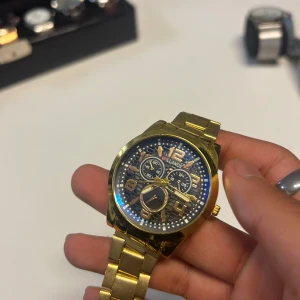 Guldig klocka från Willande - Snygg guldig klocka från Willande med länkarmband i metall och detaljerad urtavla med flera små ur. Urtavlan är svart med guldiga siffror och visare, och armbandet har klassisk länkdesign. Perfekt accessoar för dig som gillar stilrena detaljer. OBS som ni ser i bild 3 och 4 så är det en liten pinne borta som jag har tappat annars har jag bara använt den en fotal gånger då jag köpt den i Dubai, priset är inte hugget i sten