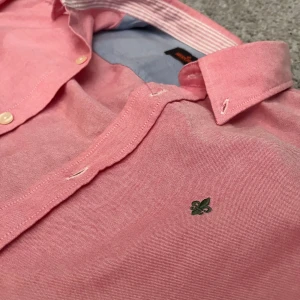 Rosa skjorta från Morris - Snygg rosa skjorta från Morris i storlek M. Skjortan har klassisk krage, vita knappar och kontrasttyg i ljusblått på insidan av manschetterna och kragen. Broderad logga på bröstet och randigt tyg på insidan av kragen. Perfekt för dig som gillar stilrena detaljer.