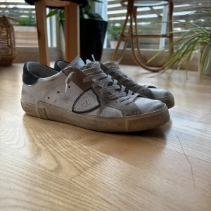 Philippe Model  - Snygga vita sneakers från Philippe Model med grå mockadetaljer och svart häl. Klassisk låg modell med rund tå, snörning och robust sula. Skorna är tillverkade i skinn och mocka, perfekta för dig som gillar en clean sommar look. Säljs pågrund av inget användande! Hör av er vid frågor köpta nya ni ser följesedeln på bild!