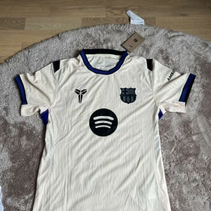 Barcelona bortatröja Nike beige - Snygg beige FC Barcelona bortatröja från Nike med svarta och blå detaljer. Tröjan har klubbmärke, sponsorlogga (Spotify) och UNHCR-tryck på ryggen. Tillverkad i lätt och ventilerande Dri-FIT-material, perfekt för fotboll eller att bära casual. Kortärmad modell med modern passform.
