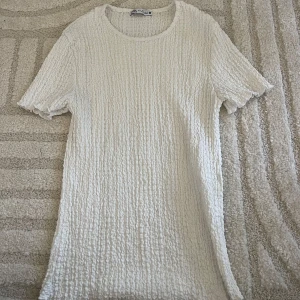 Vit ribbad t-shirt från Zara - Säljer en vit t-shirt från Zara med ribbad struktur och korta ärmar. T-shirten har en rund halsringning och är gjord i ett mjukt material som känns skönt mot huden. Perfekt för en clean och stilren look.