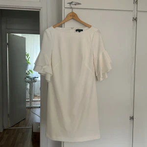 Vit volangklänning från Lauren Ralph Lauren - Superfin vit kortklänning från Lauren Ralph Lauren med volangärmar och rund halsringning. Klänningen har en stilren, rak passform och är gjord i ett mjukt polyestertyg. Perfekt för dig som gillar en clean och elegant look med extra detalj på ärmarna.