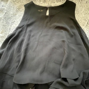 Svart ärmlös blus från Zara Basic - Säljer en svart ärmlös blus från Zara Basic i flowy modell. Toppen har en liten knappdetalj i nacken och är perfekt för dig som gillar stilrena och enkla plagg med snygg passform.