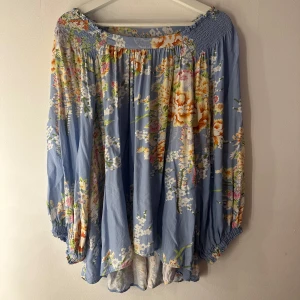 Blommig blå blus med vida ärmar - Superfin blommig blus i ljusblått med stora, färgglada blommor i vitt, orange och rosa. Vida långa ärmar med resår vid axlar och ärmslut ger en avslappnad och flowy vibe. Tillverkad i 100% viskos som känns lätt och luftig. Perfekt för vår och sommar! Storlek L men något stor i storleken 
