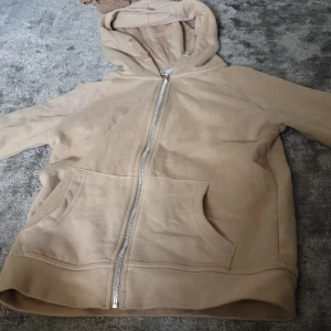 Beige hoodie med dragkedja och huva - Beige hoodie med hel dragkedja och stor ficka framtill. Tröjan har huva och är tillverkad i mjukt bomullsmaterial. Perfekt för chill dagar och enkel att matcha med jeans eller mjukisbyxor.