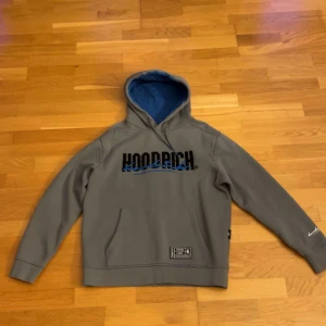 Grå hoodie från Hoodrich med blå detaljer - Snygg grå hoodie från Hoodrich med svart och blå logga på bröstet och ryggen. Tröjan har huva med blått foder, känguruficka framtill och ribbade muddar. Perfekt för dig som gillar streetwear och vill ha en stilren men ändå unik look. (byxorna finns också, går att diskutera pris)