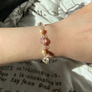 Sött armband med pärlor och hjärta - Armband med mix av pärlor och glaspärlor i ljusrosa, vitt och beige. Mittendelen har ett glittrigt rosa hjärta och små blommor i glas. Silverfärgad kedja och lås. Perfekt för dig som gillar unika och romantiska smycken.