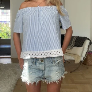 Randig offshoulder topp med spetskant - Säljer en ljusblå och vit randig offshoulder topp med korta ärmar. Toppen har en söt vit spetskant längst ner som ger en romantisk touch. Perfekt för varma dagar och enkel att styla med jeansshorts eller kjol.