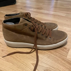 Bruna sneakers i mocka med snörning från ecco - Snygga bruna sneakers i mocka med högt skaft och vita sulor. Skorna har rund tå och klassisk snörning med matchande bruna skosnören. Perfekta för dig som gillar en stilren och avslappnad look. Priset kan diskuteras