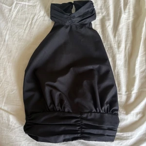 Svart halterneck topp med knytning på baksidan - Snygg svart halterneck topp med hög krage och breda knytband i ryggen. Toppen är lätt croppad med rynkade detaljer både upptill och nertill. Perfekt för en utekväll eller till en fin middag!