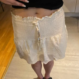 Krämvit kortkjol med snörning från Zara - Supersöt krämvit kortkjol från Zara med smockad midja och volangkant upptill. Kjolen har knytband med pärldetaljer och tofsar framtill. Perfekt för sommarens alla tillfällen och lätt att matcha med crop tops eller linnen.