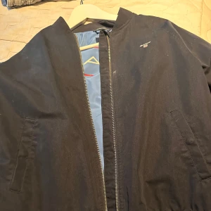 Svart bomberjacka från Gant - Snygg svart bomberjacka från Gant med klassisk passform. Jackan har dragkedja framtill, två fickor och blått innerfoder med stor Gant-logga. Perfekt för dig som vill ha en stilren och tidlös look.