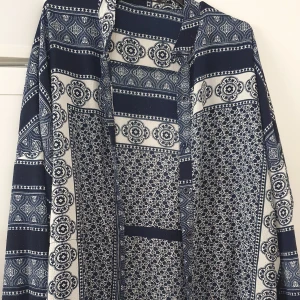 Klänning - Säljer en lång, blå och vit klänning från Boohoo med detaljerat mönster. Klänningen har lång ärm och öppen front, perfekt för att ge outfiten ett lyft med sitt unika tryck.