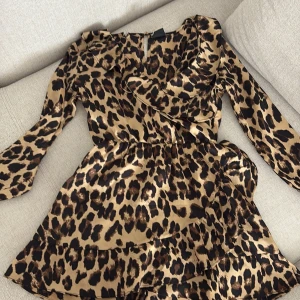 Leopardmönstrad kort klänning med volang - Snygg kort klänning med leopardmönster i beige och brunt. Klänningen har långa ärmar, volangdetaljer vid halsringningen och kjolen, samt en lätt glansig satinliknande yta. Perfekt för dig som vill sticka ut med en trendig look.