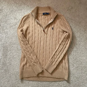 Beige stickad tröja Polo Ralph Lauren - Kabelstickad beige tröja från Polo Ralph Lauren med half zip och klassisk broderad logga på bröstet. Tröjan har hög krage, långa ärmar och ribbade muddar. Perfekt för lager-på-lager och snygg till jeans eller chinos. (Pris kan diskuteras)