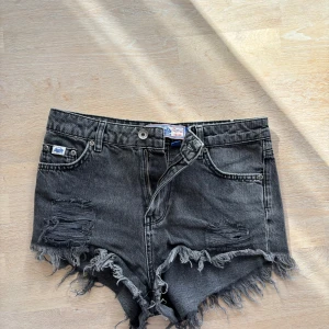 Svarta jeansshorts från Superdry - Riktigt snygga svarta jeansshorts från Superdry med hög midja, slitna detaljer och fransig kant. Klassisk femficksmodell och cool vintagekänsla. Perfekta för en edgy sommarlook!