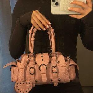 Rosa handväska med nitar och hjärtdetalj - Säljer en rosa handväska i croco-mönstrat syntetmaterial med coola nitar och spännen. Väskan har dubbla handtag, sidofickor och en hjärtformad tag med nitar. Perfekt för dig som vill sticka ut med en edgy och söt accessoar.