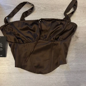 Brun satin korsettopp från NA-KD - Supersnygg brun korsettopp från NA-KD i glansigt satinmaterial. Toppen har smala axelband, markerad byst och lätt utsvängd nederkant. Perfekt för att lyfta vilken outfit som helst med sin trendiga look och lyxiga känsla.