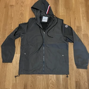 Grå vindjacka från Moncler med huva - Snygg grå vindjacka från Moncler med huva och dragkedja framtill. Jackan har Moncler-logga på ärmen, svarta snören och en cool detalj med rödvit rand på huvan. Perfekt för dig som vill ha en stilren och sportig look. Oanvänd, pris kan diskuteras. Strlk 1/S