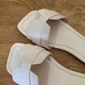 Beiga sandaler med fyrkantig tå - Snygga beiga sandaler med breda korsade remmar och fyrkantig tå. Platt sula och öppen design, perfekta för varma dagar. Tillverkade i glansigt syntetmaterial som ger en modern look.