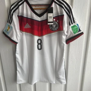 Tysklands Özil #8 VM-tröja Adidas - Säljer en officiell Tyskland fotbollströja från VM med Özil och nummer 8 på ryggen. Tröjan är vit med svarta och röda detaljer, Adidas-logga, landslagsemblem och FIFA-patch på ärmen. Tillverkad i lätt funktionsmaterial med korta ärmar och klassisk V-ringning.