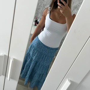 Blå blommig långkjol - Supersöt blå långkjol med småblommigt mönster i blått och grönt. Kjolen har volangdetaljer och resår i midjan för en bekväm passform. Luftig och fin och når ner till fotleden på mig som är 166cm. Vid frågor hör av dig!