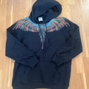 Marcelo Burlon svart hoodie med fjädertryck - Svart hoodie från Marcelo Burlon County of Milan med ikoniskt färgglatt fjädertryck över axlar och bröst. Tröjan har huva med snörning, känguruficka och är tillverkad i 100% bomull. Perfekt statement-plagg för dig som vill sticka ut.