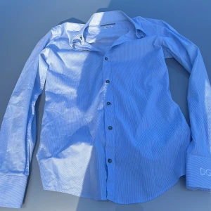 Blå randig skjorta från Dolce & Gabbana - Snygg blå skjorta från Dolce & Gabbana Gold med tunna vita ränder, broderad DG-logga på manschetten och svarta knappar med märkets namn. Klassisk krage och lång ärm, perfekt för dig som vill ha en stilren och exklusiv look.