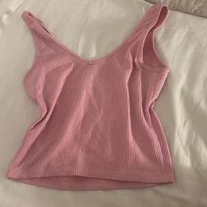 Rosa ribbad linnetopp med v-ringning - Säljer en söt rosa linnetopp med smala vita ränder och v-ringad hals. Toppen är ribbad och ärmlös, perfekt för varma dagar. Den har en enkel och snygg design som är lätt att matcha med jeans eller kjol.