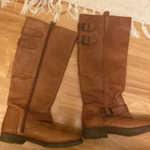 Bruna skinnstövlar Tommy Hilfiger - Snygga bruna stövlar från Tommy Hilfiger Denim. Höga med två spännen upptill och ett vid ankeln, grov sula och dragkedja på insidan. Perfekta till höst- och vinterlooken, med klassisk logga på sidan och metallmärke upptill.