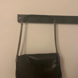 Svart axelväska med kedjerem - Snygg svart axelväska i skinn med silverfärgad kedjerem. Väskan har lock med magnetknäppning och ett stort huvudfack med dragkedja samt flera innerfack. Perfekt storlek för det viktigaste och ger en cool vibe till din outfit.