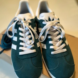 Adidas Gazelle mörkgröna sneakers - Snygga Adidas Gazelle sneakers i mörkgrön mocka. Skorna har vit plös och häl med Adidas-logga, samt vita snören och extra gröna skosnören.  Använda 2 dagar. Hund finns i hemmet. Något små i storlek.