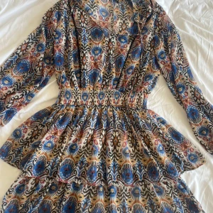 Mönstrad kortklänning från Zara XS - Färgstark kortklänning från Zara i storlek XS med unikt mönster i blått, beige, svart och rost. Klänningen har lång ärm, markerad midja med smock och volangig kjol. Tunt, luftigt material som passar perfekt för varmare dagar.