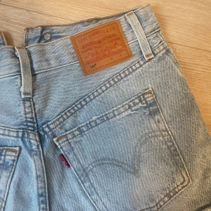 Levi's ljusblå jeansshorts 501 - Säljer ett par klassiska ljusblå jeansshorts från Levi's modell 501. De är högmidjade med råa, fransiga kanter och har fem fickor samt knappar framtill. Perfekta för sommaren och ger en avslappnad vintagekänsla. Ikonisk läderpatch bak och röd Levi's-tagg på fickan.