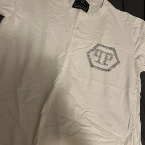 Vit t-shirt från Philipp Plein med strass - Vit t-shirt från Philipp Plein med korta ärmar och rund hals. På bröstet sitter ett stort PP-logo i silverfärgade strass-stenar som ger en lyxig vibe. Tillverkad i mjuk bomull för skön känsla och snygg passform.