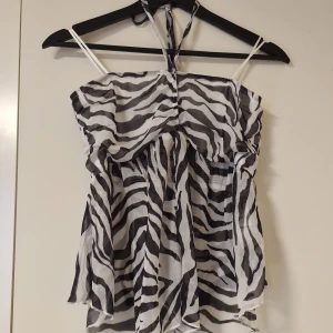 Zebramönstrad ärmlös linnetopp - Snygg ärmlös linnetopp med zebramönster i svart och vitt. Toppen har tunna axelband och knyt i nacken för en cool halterneck-look. Luftigt material och volangig nederdel ger en skön och trendig vibe, perfekt för varma dagar.