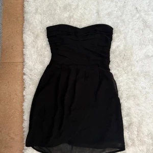 Svart axelbandslös kortklänning - Snygg svart axelbandslös kortklänning med draperad överdel och omlottkjol. Perfekt för dig som vill ha en stilren och elegant look. Klänningen har en figurnära passform och är tillverkad i ett lätt, flowy material.