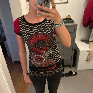 Desigual mönstrad t-shirt i rött och svart - Unik t-shirt från Desigual med coola grafiska mönster i svart, rött och grått. Framsidan har vågiga och blommiga tryck, baksidan är rutig och ärmarna är korta med röda detaljer. Figurnära passform och mjukt bomullsmaterial.