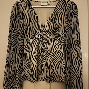 Zebramönstrad blus från Vila - Säljer en snygg zebramönstrad blus från Vila i svart och vitt. Blusen har v-ringning, långa ärmar och lätt puff vid axlarna. Materialet är tunt och flowy, perfekt för att sticka ut med ett coolt mönster. Passformen är normal och modellen är lite lös nertill.