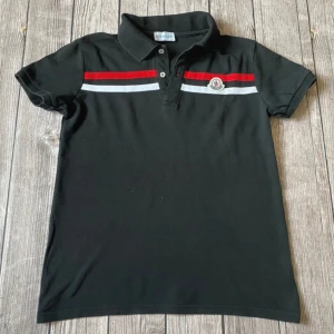 Svart pikétröja Moncler med röd detalj - Svart pikétröja från Moncler med klassisk krage och knappar framtill. Framsidan har röda och vita ränder över bröstet samt Moncler-logga. Baksidan av kragen har stor röd Moncler-text. Kortärmad och i mjuk bomull, perfekt för en clean och sportig look.