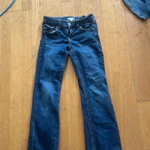 Blå bootcut jeans med fickdetaljer - Snygga blå jeans med bootcut passform och coola fickdetaljer både fram och bak. Klassisk denim med kontrastsömmar och knappar på bakfickorna. Perfekta för dig som gillar en avslappnad men ändå trendig look. Dom håller på att få ett liten hål där bak vid fickan.
