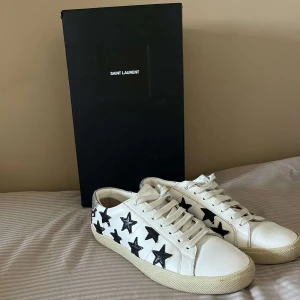 Saint Laurent sneakers med stjärnor - Snygga vita Saint Laurent skor i skinn med svarta stjärnor, vita snören och beige sula. Perfekt för dig som vill ha moderna och snygga skor till skolstarten. Skorna är i storlek 40 men passar större. Nypris: cirka 7000kr, mitt pris: 1850kr. Hör av er vid minsta lilla fråga. 