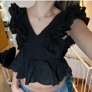 Svart volangblus med v-ringning - Supersnygg svart blus med djup v-ringning och massor av volanger längs axlar och bröst. Normal i storleken