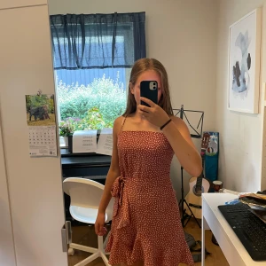 Rosa klänning  - Superfin rosa kortklänning från Sundae Muse med vita prickarliknande former, volang och knyt i midjan. Klänningen har smala axelband och dragkedja i ryggen. Perfekt för sommarens alla tillfällen!