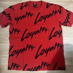 Röd t-shirt med svart print  - Röd t-shirt från Squad med svart textprint 'Loyalty' över hela plagget. Klassisk rund halsringning och korta ärmar. Skön passform och tillverkad i mjuk bomull, perfekt för en streetwear-look.