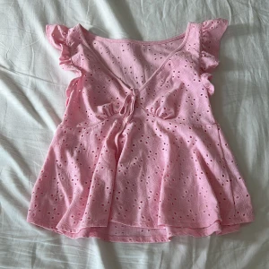 Rosa blus med volangärmar och knyt - Supersöt ljusrosa blus med broderade hålmönster, volangärmar och v-ringning med knyt framtill. perfekt för varma dagar. 