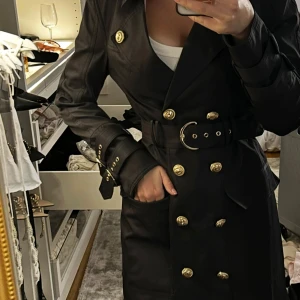 Svart trenchcoat med guldknappar  - Svart kappa från Coventina Collection som tyvärr inte kommer till användning för mig.