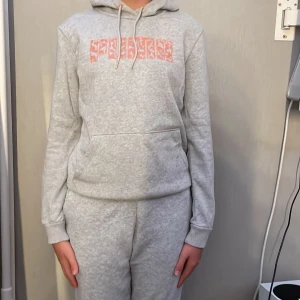 Grå hoodie från Puma med orange tryck - Säljer en grå hoodie från Puma med stort orange logotyp-tryck framtill. Tröjan har huva med snörning och en stor magficka. Mjuk och skön bomullsblandning, perfekt för chill eller träning. Långärmad och normal passform.