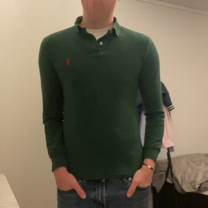 Grön pikétröja Polo Ralph Lauren XS - Mörkgrön långärmad pikétröja från Polo Ralph Lauren i custom slim fit. Klassisk krage, knäppning vid halsen och röd broderad logga på bröstet. Tillverkad i 100% bomull, perfekt för en clean och stilren look.